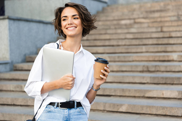 Photo of young cute beautiful woman walking outdoors holding laptop computer holding coffee. - Valokuva, kuva