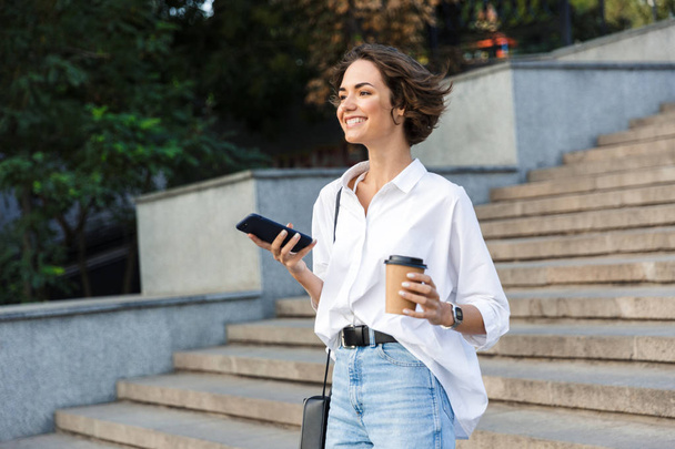 Photo of young cute beautiful woman walking on the street using mobile phone holding coffee. - Photo, Image