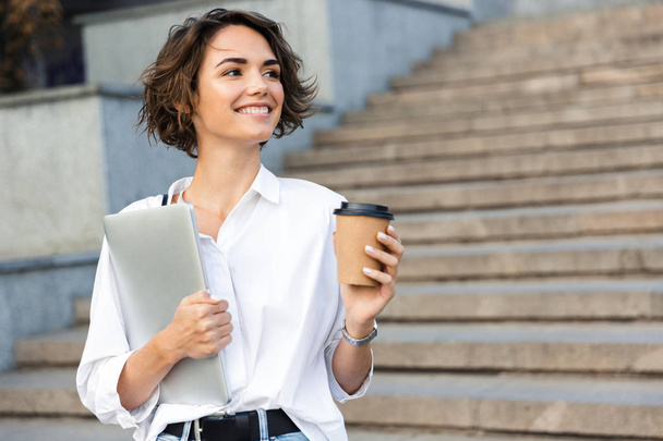Image of young cheerful beautiful woman walking outdoors holding laptop computer holding coffee. - Foto, Bild