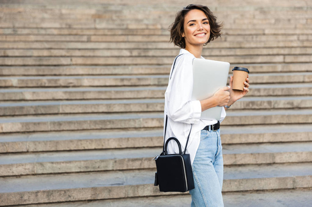 Εικόνα του νεαρό χαρούμενα όμορφη γυναίκα που περπατά κρατώντας σε εξωτερικούς χώρους φορητό υπολογιστή κρατώντας καφέ. - Φωτογραφία, εικόνα