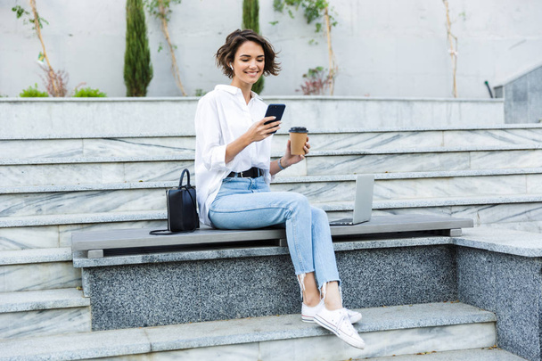 Belle jeune femme dans les écouteurs assis à l'extérieur dans la rue, parlant sur un téléphone portable, buvant du café
 - Photo, image