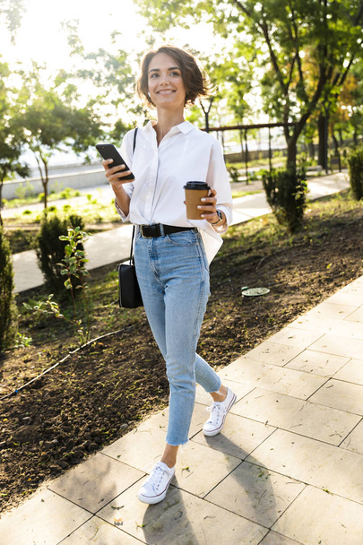 Preciosa mujer joven caminando por la calle, sosteniendo el teléfono móvil, bebiendo café
 - Foto, imagen