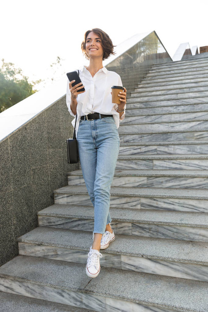 Cheerful young woman walking downstairs at the street, using mobile phone, drinking coffee - Фото, изображение