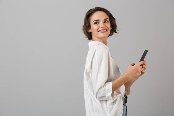 Image of happy young business woman posing isolated over grey wall background using mobile phone chatting. - Photo, Image