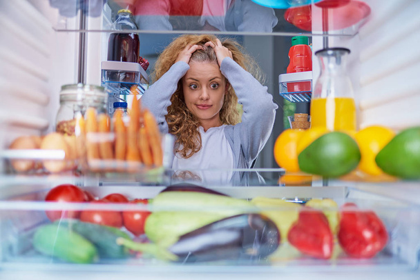 Confused woman choosing what to eat. Hands on head. Picture taken from the inside of fridge full of groceries. - Photo, Image