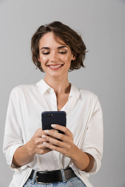 Image of happy young business woman posing isolated over grey wall background using mobile phone chatting. - Foto, Imagem