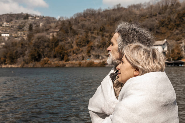 beautiful mature couple relaxing sitting on the dock wrapped in a blanket on a winter day - Φωτογραφία, εικόνα
