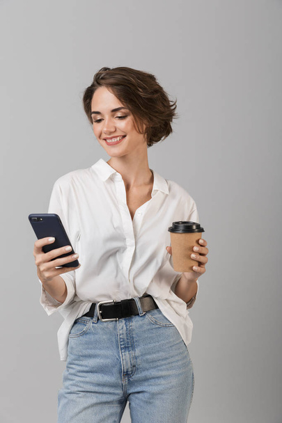 Image of happy young business woman posing isolated over grey wall background chatting by mobile phone drinking coffee. - Foto, afbeelding