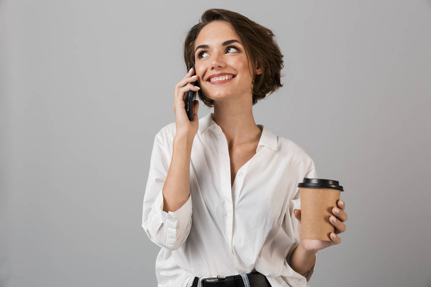 Image of happy young business woman posing isolated over grey wall background talking by mobile phone drinking coffee. - Фото, зображення