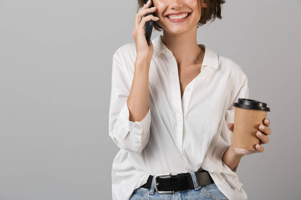 Cropped photo of happy young business woman posing isolated over grey wall background talking by mobile phone drinking coffee. - Фото, изображение