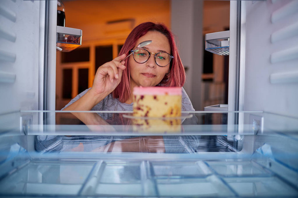 Žena jíst dort stoje před z lednice. Obrázek převzat z vnitřku lednice. - Fotografie, Obrázek