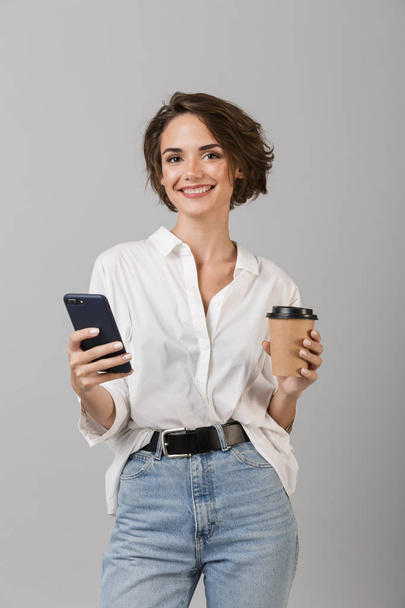 Image of happy young business woman posing isolated over grey wall background chatting by mobile phone drinking coffee. - Valokuva, kuva
