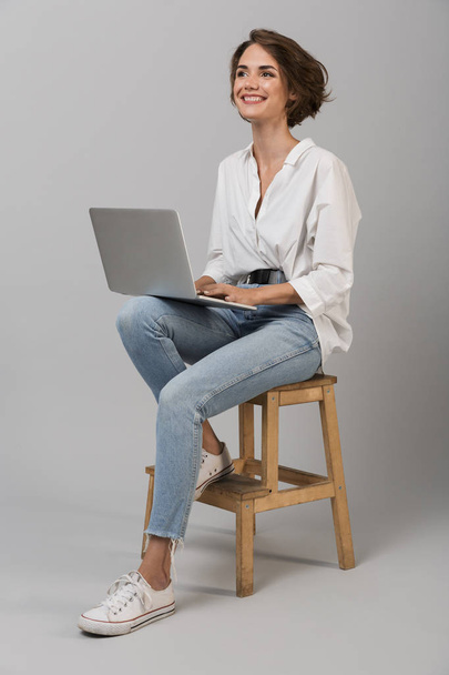 Image of young business woman posing isolated over grey wall background sitting on stool using laptop computer. - Fotografie, Obrázek