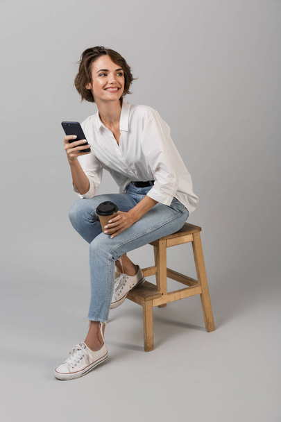 Podobu mladé ženy představují samostatný šedá stěna pozadí sedí na stoličce pomocí přenosného počítače chatování telefonicky. - Fotografie, Obrázek