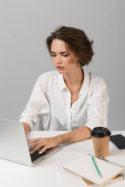 Изображение молодой серьезной деловой женщины, позирующей на фоне серой стены, сидящей за столом с помощью ноутбука
. - Фото, изображение