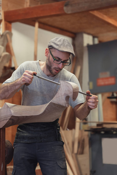 Carpintero trabajando en artículo de madera en taller, tomando medidas. Retrato de perfil de medio cuerpo de un hombre con gafas, suéter y gorra, con fondo borroso y espacio para copiar
. - Foto, Imagen