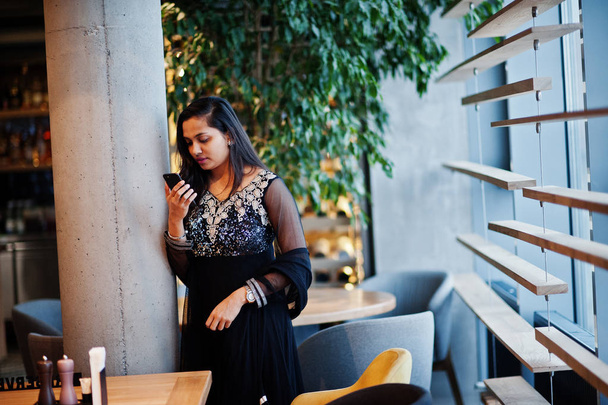 Pretty indian girl in black saree dress posed at restaurant with mobile phone at hand. - Фото, зображення