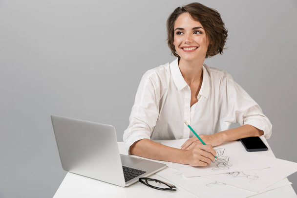 Image of young business woman posing isolated over grey wall background sitting at the table using laptop writing notes. - Fotoğraf, Görsel