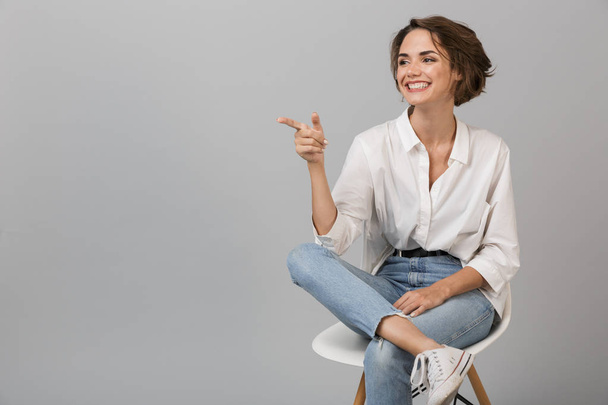 Image of young business woman posing isolated over grey wall background sitting on stool pointing. - Fotoğraf, Görsel