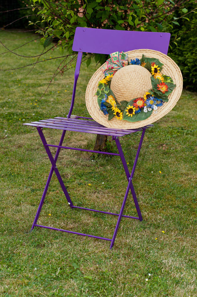 Uma velha cadeira dobrável lilás com um chapéu de palha de abas largas. No chapéu há algumas flores
 - Foto, Imagem
