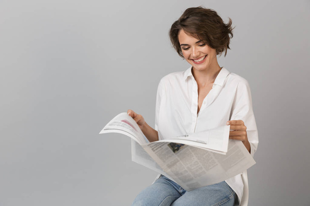 Photo of a happy young woman sitting on stool isolated over grey background holding newspaper reading. - Photo, Image