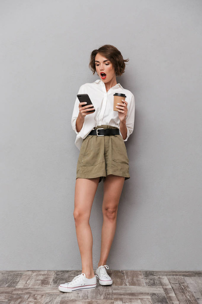 Full length image of excited european woman 20s holding takeaway coffee and using smartphone isolated over gray background - Фото, изображение