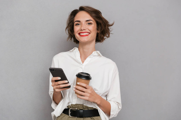 Kuva tyytyväinen nainen 20s tilalla takeaway kahvia ja matkapuhelimen eristetty harmaalla pohjalla
 - Valokuva, kuva