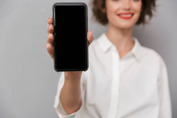 Mladá žena 20s s mobilním telefonem a zobrazení copyspace obrazovky nad šedým pozadím, samostatný - Fotografie, Obrázek