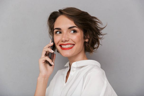 Фото милої жінки 20-х посміхається і говорить на мобільному телефоні ізольовано на сірому фоні
 - Фото, зображення