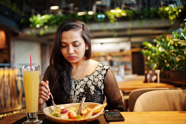 Pretty indian girl in black saree dress posed at restaurant, sitting at table with juice and salad. - Φωτογραφία, εικόνα