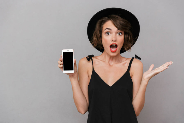 Photo of beautiful woman 20s wearing black dress and hat holding smartphone isolated over gray background - Foto, afbeelding