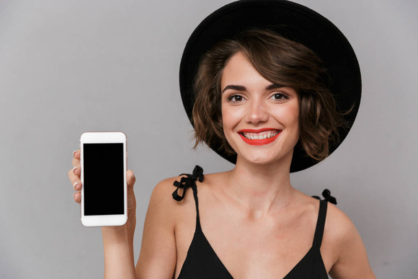 Photo of gorgeous woman 20s wearing black dress and hat holding smartphone isolated over gray background - 写真・画像