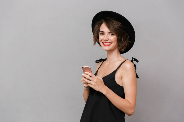 Photo of beautiful woman 20s wearing black dress and hat smiling at camera while holding smartphone isolated over gray background - Foto, Imagen
