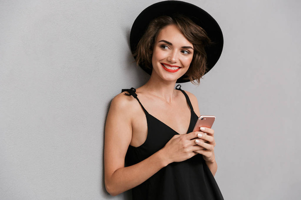 Photo of attractive woman 20s wearing black dress and hat smiling at camera while holding smartphone isolated over gray background - Foto, Imagen