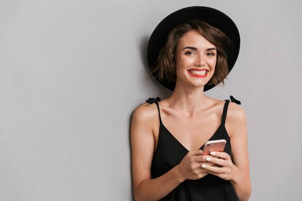 Photo of joyful woman 20s wearing black dress and hat smiling at camera while holding smartphone isolated over gray background - Фото, изображение