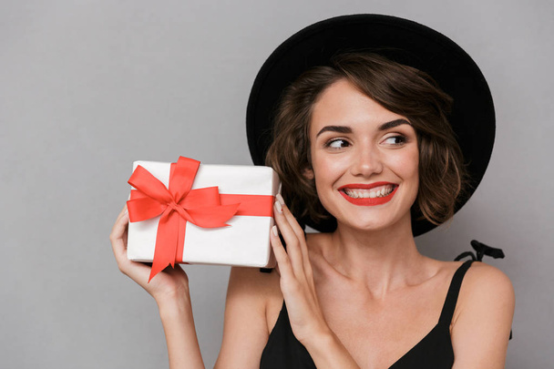 Photo of stylish woman 20s wearing black dress and hat holding present box isolated over gray background - Foto, Imagen
