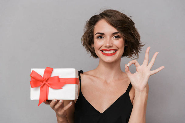 Photo of gorgeous woman 20s wearing black dress holding gift box isolated over gray background - Φωτογραφία, εικόνα