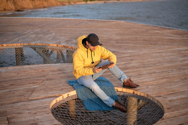 hombre joven en cortavientos amarillo y gorra negra se sienta en el muelle de madera, utiliza tableta
 - Foto, imagen