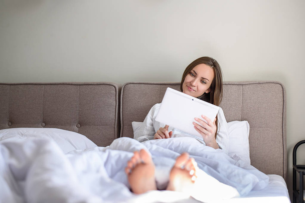 Mujer acostada en la cama bajo la manta en la tableta, navegación por Internet, noticias, buenos días
. - Foto, Imagen