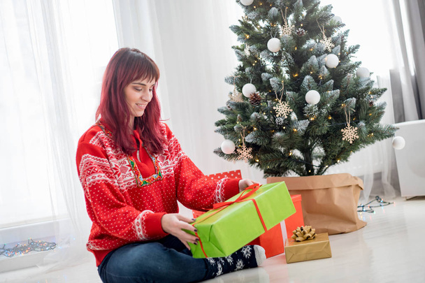 Happy young woman sitting on white floor near christmas tree holding gift box - Photo, Image