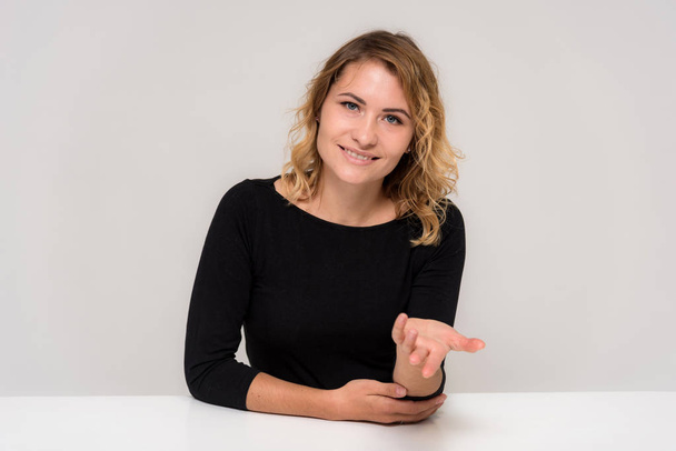 Portrait of a beautiful blonde girl on a white background at the table. She is sitting right in front of the camera, smiling and looking happy. - Foto, immagini