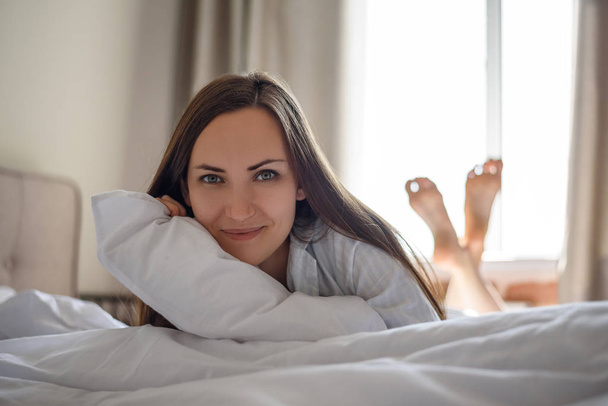 Frontal portrait of brunette girl in bed with white sheet and pillow. - Фото, изображение