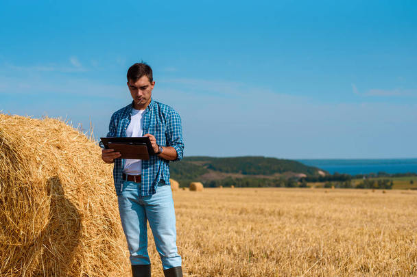 Vista frontal del agricultor agrónomo masculino con una tableta en jeans azules y camisa y camiseta blanca en el campo con pajar
 - Foto, Imagen