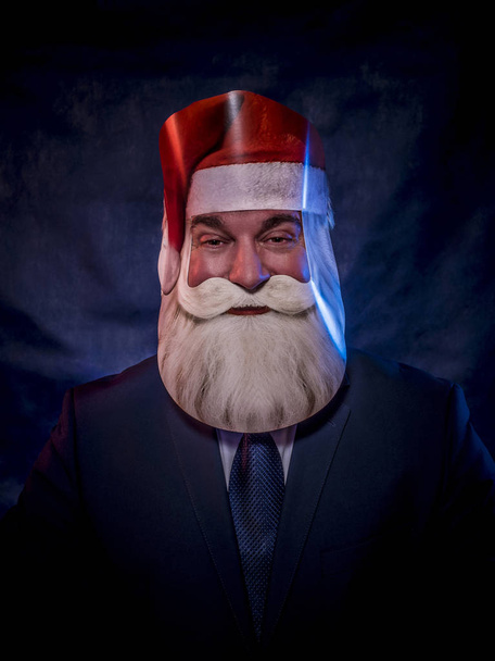 A man in a dark suit is playing the role of Santa Claus in a cardboard mask of Santa Clausagainst a blue background. - 写真・画像