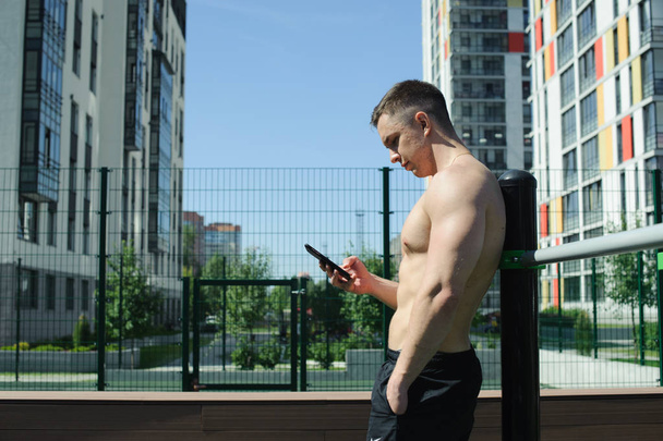 Guapo sexy hombre culturista atleta hombre mirando a la pantalla del teléfono inteligente en la mañana soleada al aire libre
. - Foto, Imagen