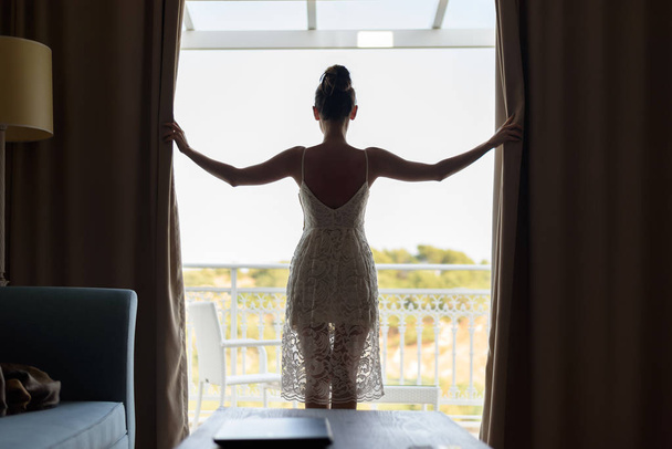Menina atraente esbelta em um vestido branco das costas fica na janela panorâmica abrindo as cortinas
. - Foto, Imagem