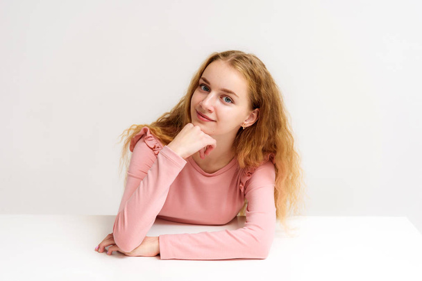 Studio portrait of a beautiful girl blonde teenager on a white background sitting at the table. She is sitting with different emotions right in front of the camera, smiling and looking happy. - Fotó, kép