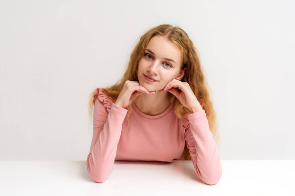 Studio portrait of a beautiful girl blonde teenager on a white background sitting at the table. She is sitting with different emotions right in front of the camera, smiling and looking happy. - Photo, image