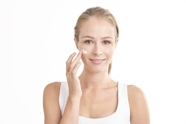 Beautiful young woman applying face cream and looking at camera while standing at isolated white background. - Φωτογραφία, εικόνα