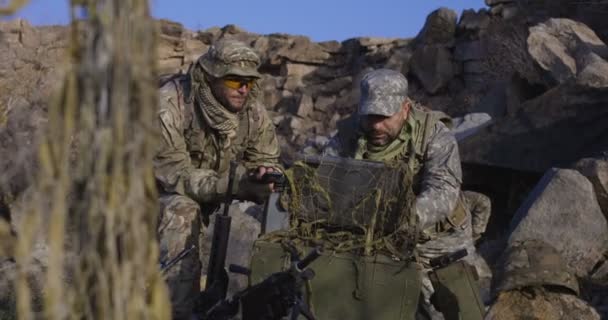 Middellange schot van volledig uitgerust en gewapende soldaten kijken naar een computer en een ander kijken naar een handbediend apparaat in het veld - Video